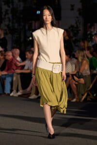 Model walking a fashion show in a sleeveless cream top and olive draped midi skirt with a wide white belt.