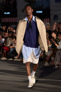 Male model walks the runway in a beige trench draped over shoulders, navy top, light blue shirt layers, and white lace-up shoes, audience in background.