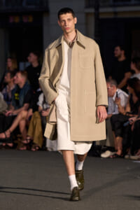 Male model walking a runway in a beige trench coat over a white outfit, paired with brown shoes and white socks.