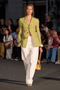 Model walking runway in a lime blazer and wide white trousers, carrying a tan handbag.