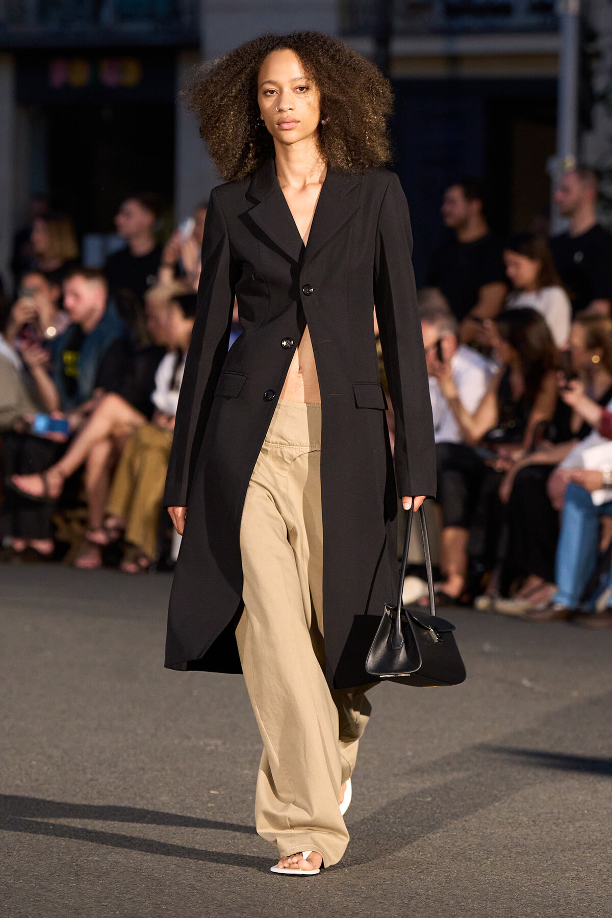 Female model walks a runway wearing a long black blazer over beige wide-leg pants, carrying a black bag; audience blurred in background.