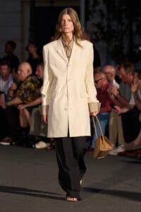 Runway model wearing a cream tailored blazer over a light shirt, black wide-leg trousers, beige sandals, and carrying a tan handbag.