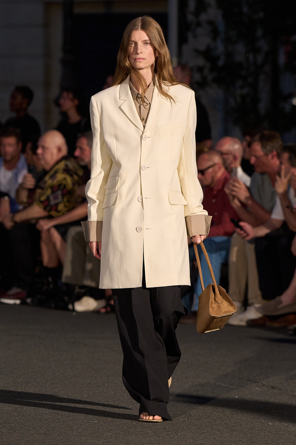 Runway model wearing a cream tailored blazer over a light shirt, black wide-leg trousers, beige sandals, and carrying a tan handbag.
