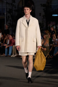 Male model on a runway wearing a cream oversized coat with matching shorts, holding a tan tote bag.