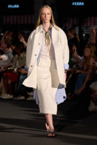 Model walking a runway in a cream oversized coat over a light blue striped shirt and beige skirt, with a green scarf at the neck.