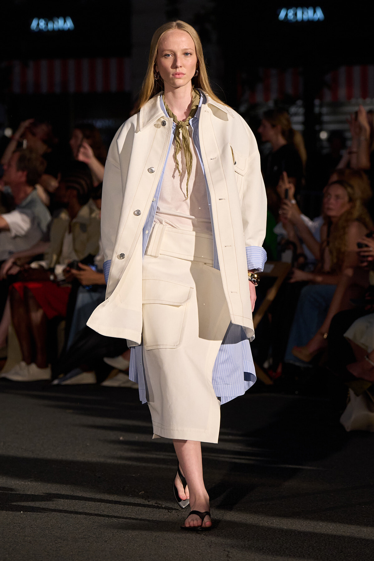 Model walking a runway in a cream oversized coat over a light blue striped shirt and beige skirt, with a green scarf at the neck.
