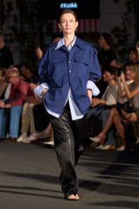 Model walks a runway in an oversized navy blue button-down shirt over a light striped shirt and black satin pants, audience watching in background.