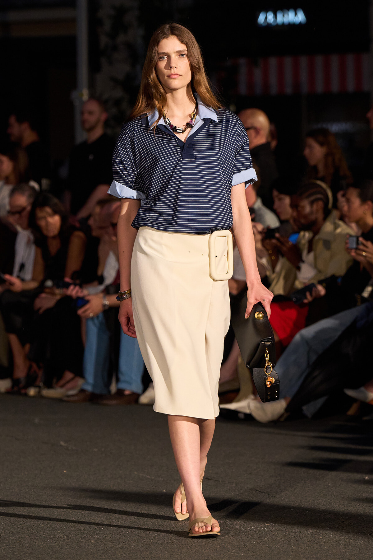 Female model walks a runway in a navy striped polo tucked into a cream midi skirt with an oversized belt; carries a black clutch with gold hardware, wearing beige sandals.