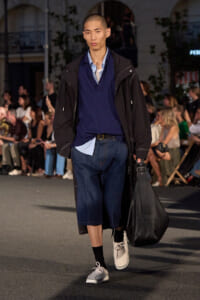 Male model on runway wearing a navy layered shirt, long black coat, knee‑length denim shorts, and a large black bag.
