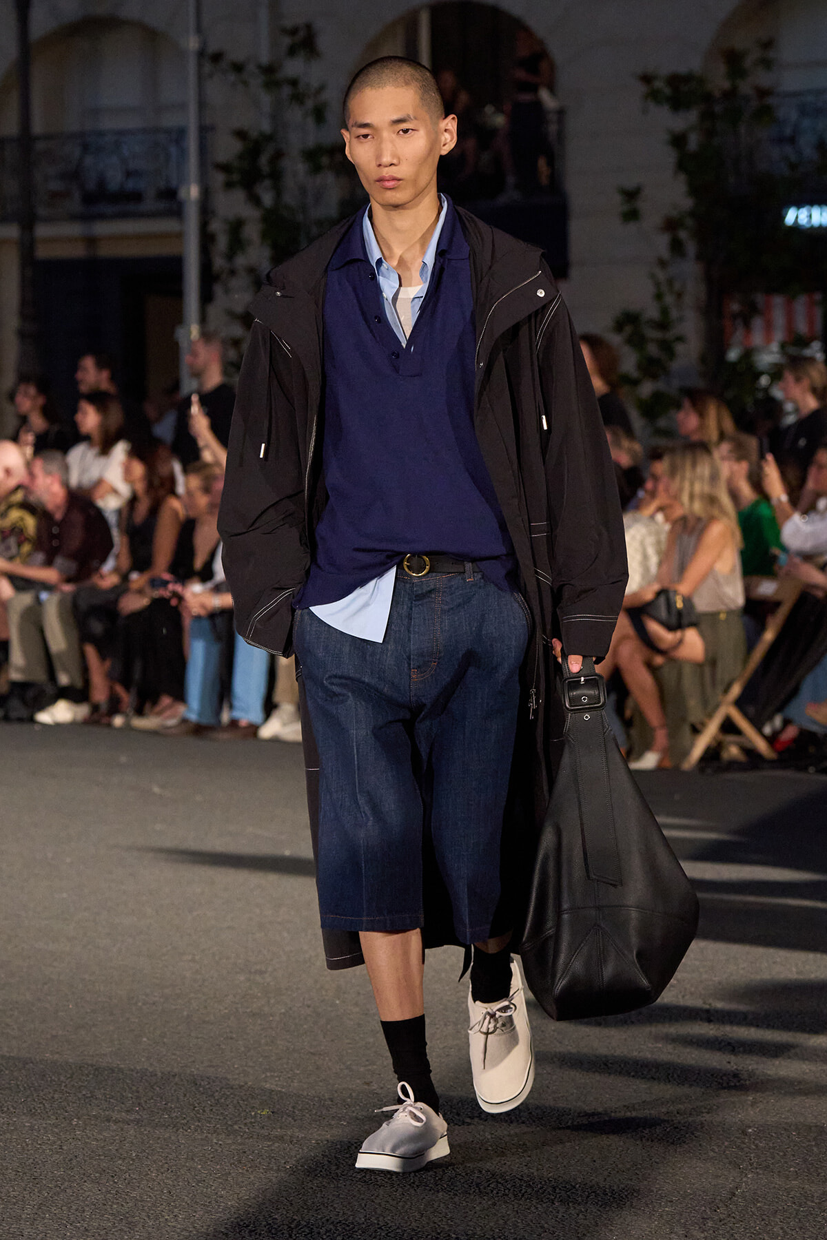 Male model on runway wearing a navy layered shirt, long black coat, knee‑length denim shorts, and a large black bag.
