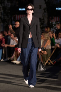 Model on a runway wearing a black blazer, wide-leg denim jeans, white pointed-toe shoes, dark sunglasses, and a black handbag.