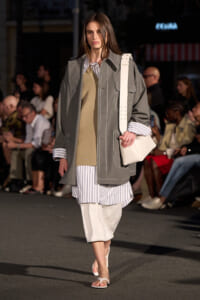 Model walks the runway in an oversized gray coat with white stitching, striped shirt underneath, and a white shoulder bag.