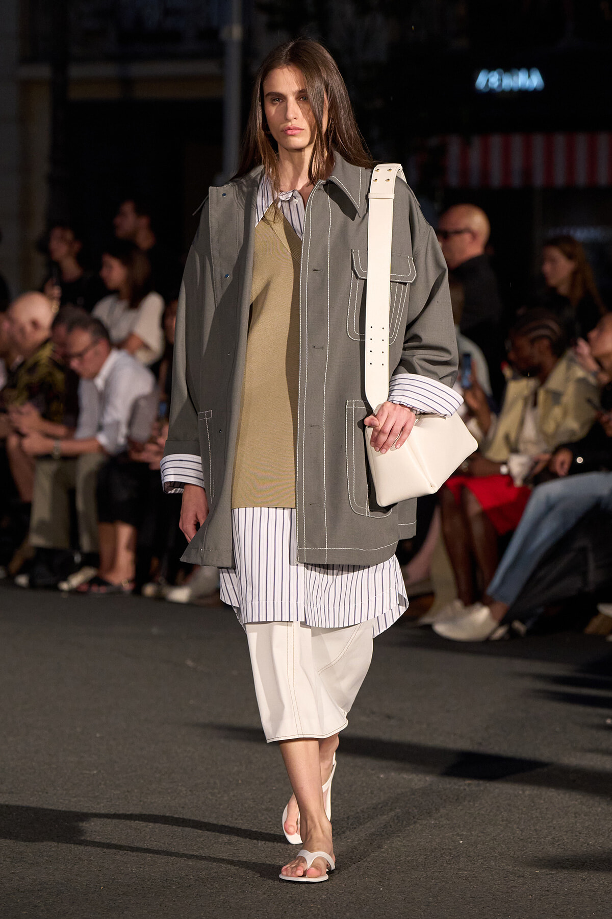 Model walks the runway in an oversized gray coat with white stitching, striped shirt underneath, and a white shoulder bag.