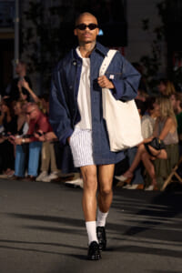 Model walking a fashion show in a dark denim oversized coat, white tee, striped shorts, white socks, black shoes, and a large white tote bag over the shoulder; audience in background.