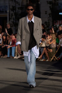 Male model on a runway wearing an oversized gray double-breasted blazer over a black sweater and blue-striped shirt, with light-washed jeans and dark sunglasses.