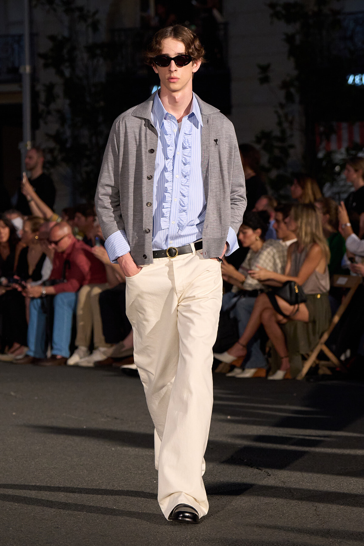 Male model on a runway wearing a light blue ruffled shirt, gray cardigan, cream wide-leg pants, black belt and sunglasses.