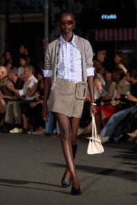 Model walks runway in a light blue ruffled shirt, gray cardigan, and beige suede mini skirt with a large belt pouch, pairing black heels and red-tinted sunglasses, carrying a small white handbag.