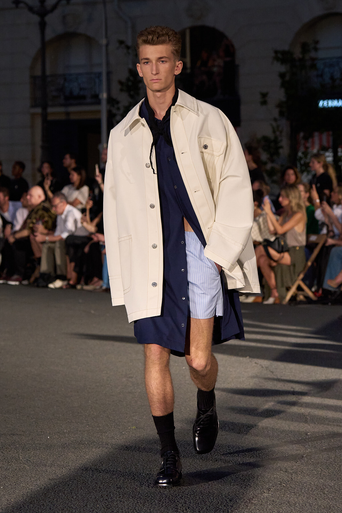 Male model walking the runway in an oversized cream jacket over a navy shirt and light blue striped shorts.