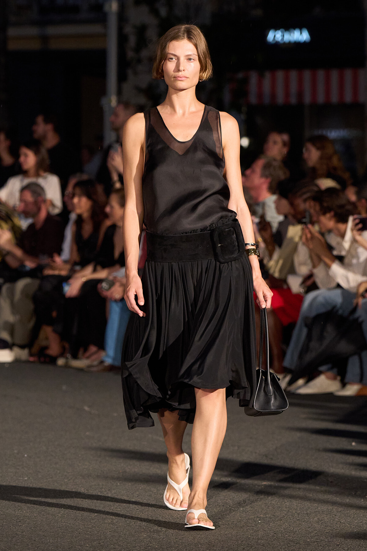 Female model walking a fashion runway in a black sleeveless satin top and voluminous black skirt, carrying a small black bag.