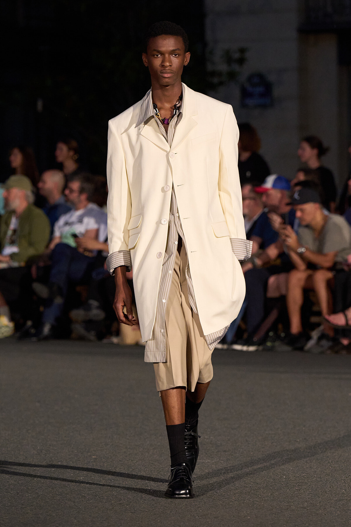 Male model on a runway wearing a cream oversized blazer over a striped shirt and beige knee-length shorts; black boots.