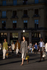 Street fashion show at dusk with models walking the runway on a city street, wearing tailored coats and carrying handbags against a grand building backdrop.