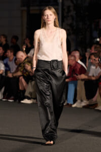 Model walks a runway in a sheer cream tank top and oversized black satin pants with a large rectangular belt. Audience in background.