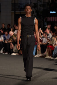 Male model walking a nighttime street-style runway in a black sleeveless top with white accents, black trousers, and a belt with a gold buckle.