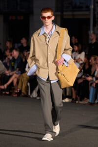Male model walking a fashion runway in a tan button-front jacket over a blue striped shirt, gray trousers, white sneakers, red sunglasses, and a large tan shoulder bag.