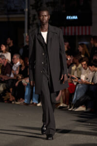 Male model walks a runway in a long black overcoat over a white tee and black suit, with spectators in the background.