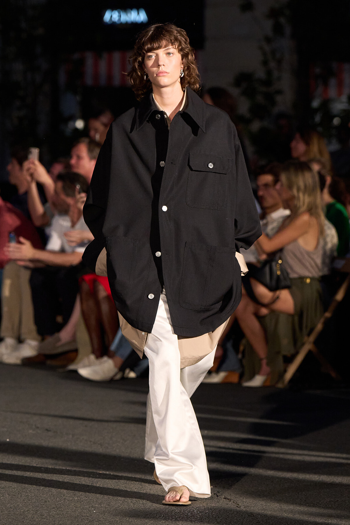 Model on a runway wearing an oversized black utility jacket over beige layers and white wide-leg pants; audience visible in background.