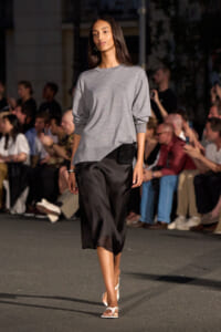 Model walks a runway in a gray oversized sweater, black satin midi skirt, white strappy sandals, and a small black waist bag. Audience visible in background.