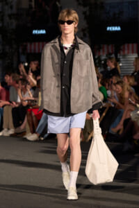 Male model on a runway wearing a gray suede jacket over a black shirt and light blue shorts, with white shoes and a large white tote bag.
