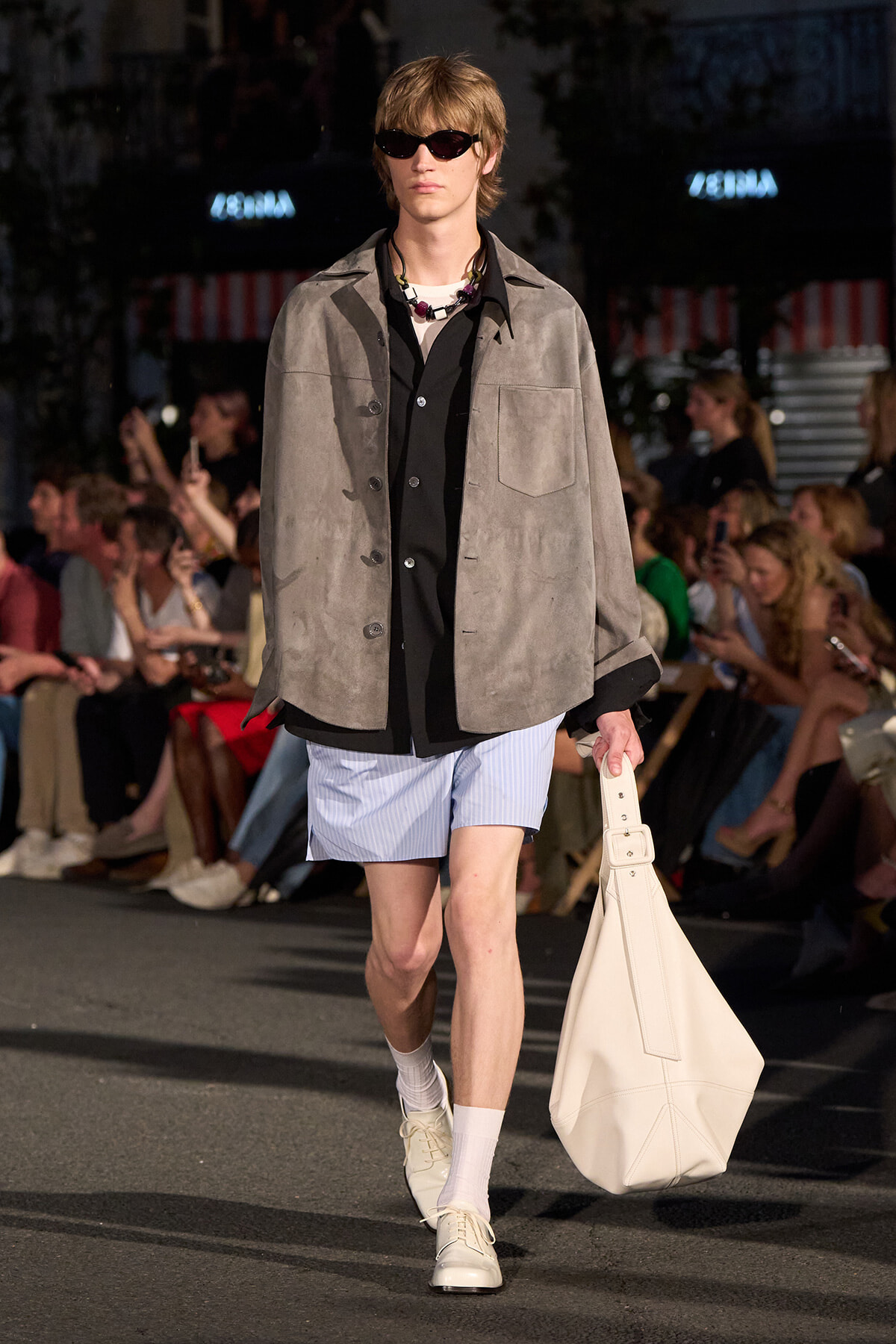 Male model on a runway wearing a gray suede jacket over a black shirt and light blue shorts, with white shoes and a large white tote bag.