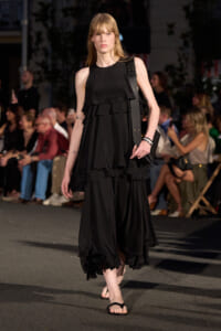 Model in a black multi-layered dress walks a runway with a black shoulder bag and flip-flop sandals; audience in background.