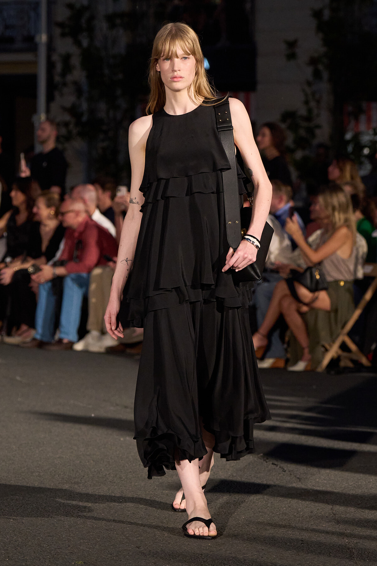 Model in a black multi-layered dress walks a runway with a black shoulder bag and flip-flop sandals; audience in background.