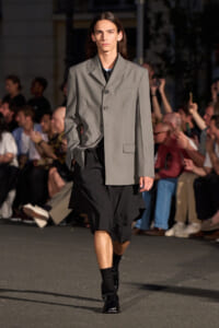 Male model walking a runway in a light gray oversized blazer over a black layered outfit, black socks and shiny black shoes, crowd in background.