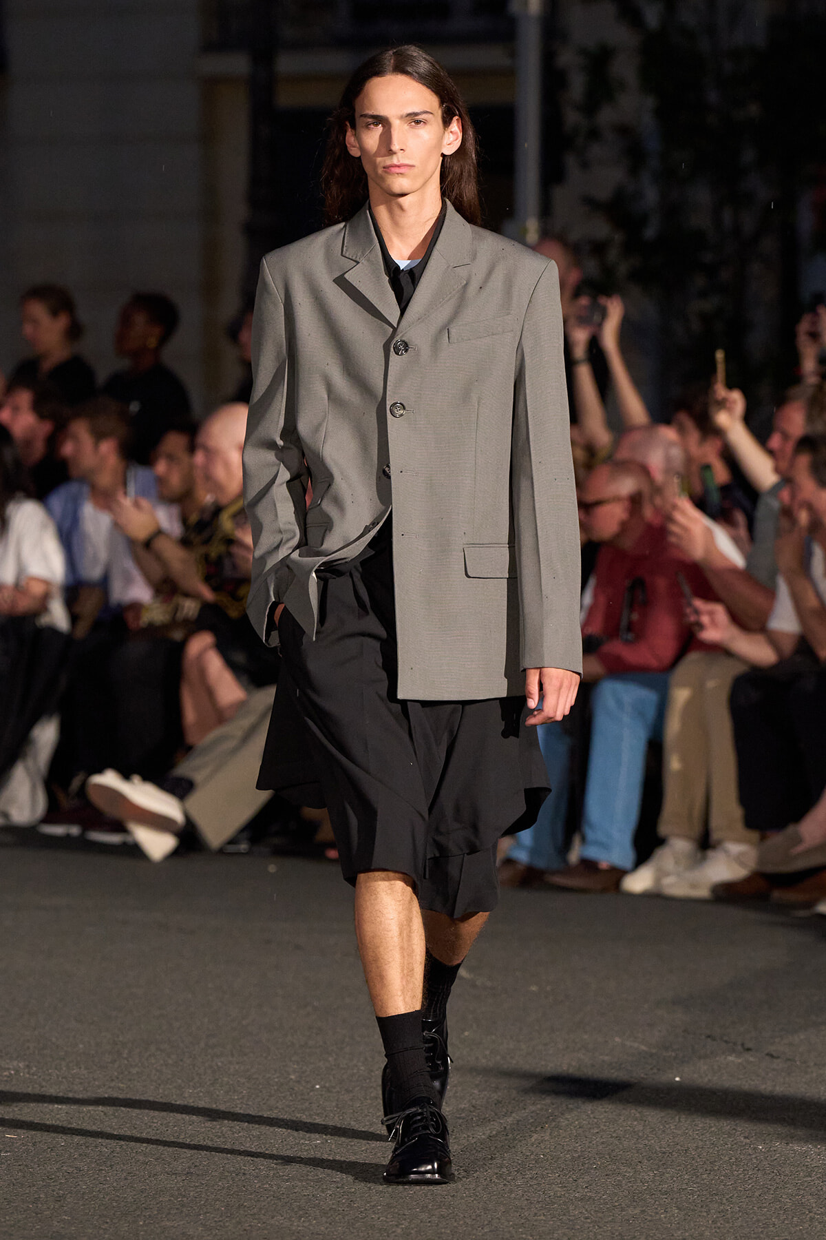 Male model walking a runway in a light gray oversized blazer over a black layered outfit, black socks and shiny black shoes, crowd in background.