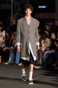 Male model walks a runway in a gray, long coat over a vertical striped shirt, black knee-length shorts, white socks, and navy sneakers while carrying a black bag; audience in background.