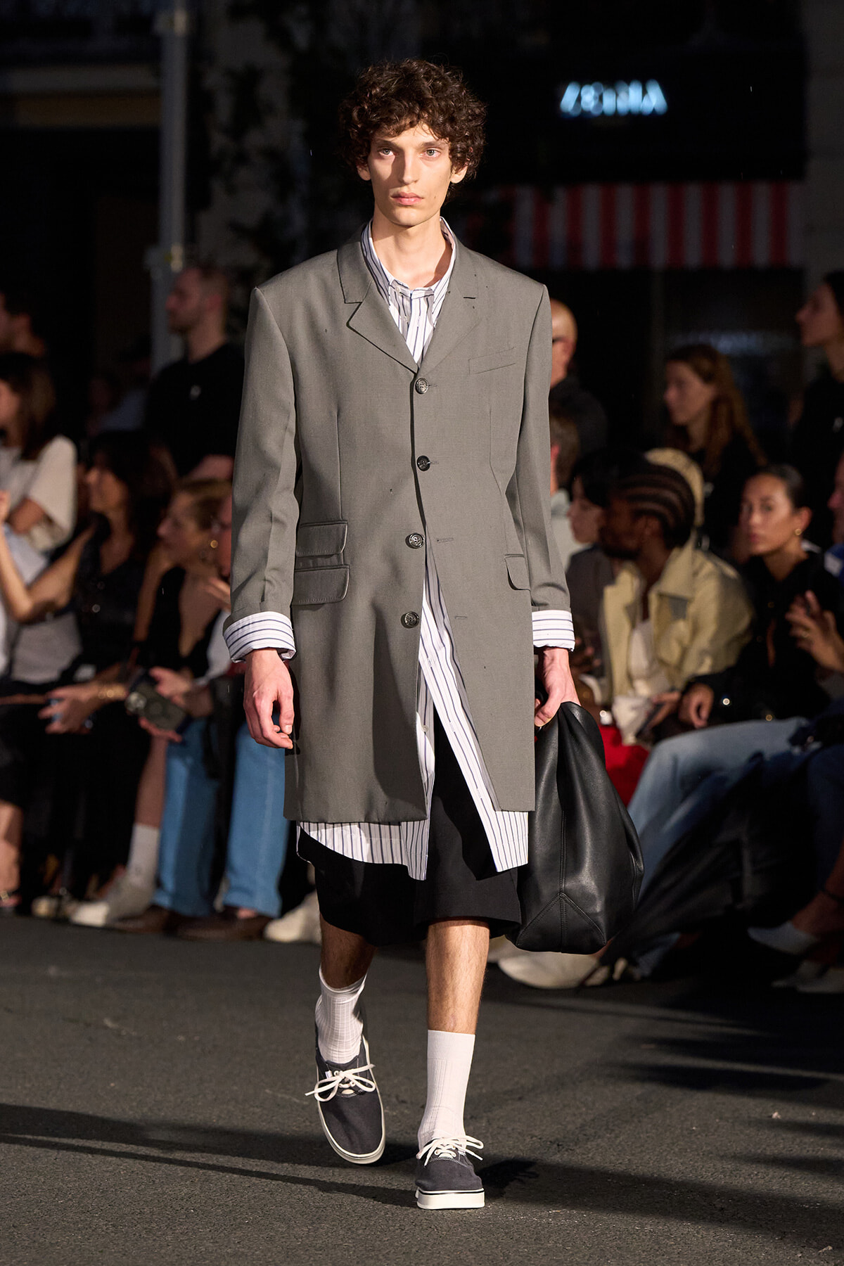 Male model walks a runway in a gray, long coat over a vertical striped shirt, black knee-length shorts, white socks, and navy sneakers while carrying a black bag; audience in background.