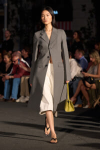 Female model walking a runway in a long gray blazer over a white midi skirt, carrying a yellow handbag.