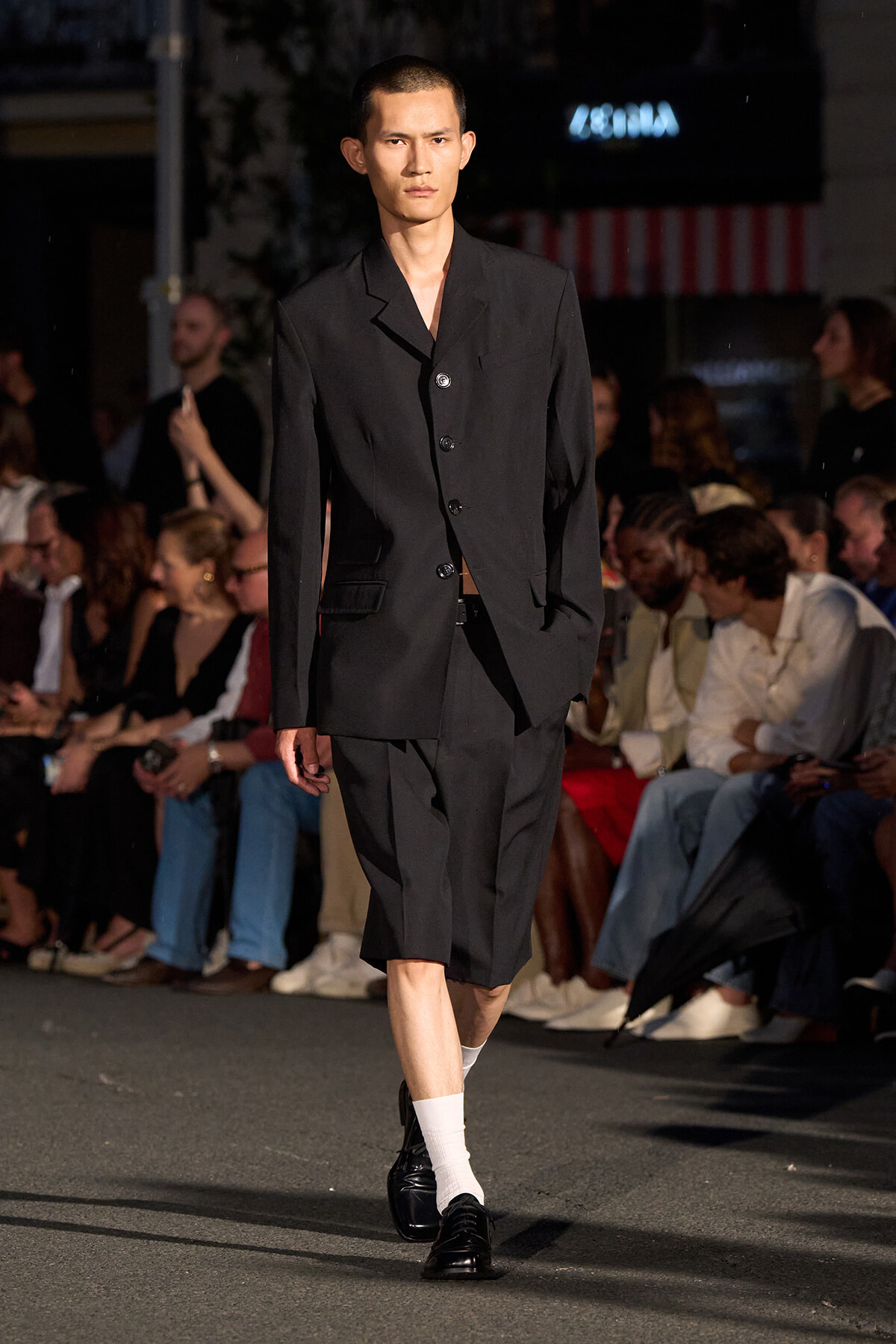 Male model walks the runway in a tailored black blazer and cropped pants; audience seated along the sides.