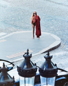 Woman in a burgundy coat and scarf standing on a circular plaza, cobblestone street in the background, wearing sunglasses and heels.