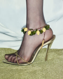Foot wearing a gold strappy stiletto with sheer black stockings and a decorative ankle bracelet featuring yellow lemon-shaped charms and green leaves.