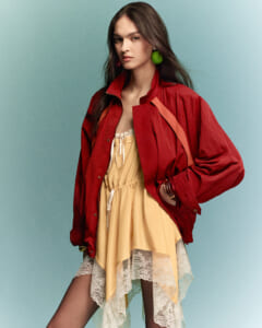 Model wearing a red jacket over a pale yellow lace-trim dress, posing against a teal background.