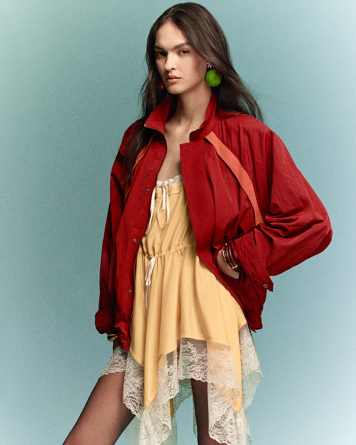 Model wearing a red jacket over a pale yellow lace-trim dress, posing against a teal background.