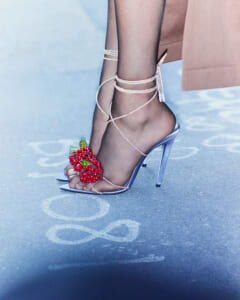 Blue metallic strappy high-heeled sandal with pearl ankle wrap and a red berry ornament on the toe, worn on a pavement with chalk markings near a pink garment surface and coat.