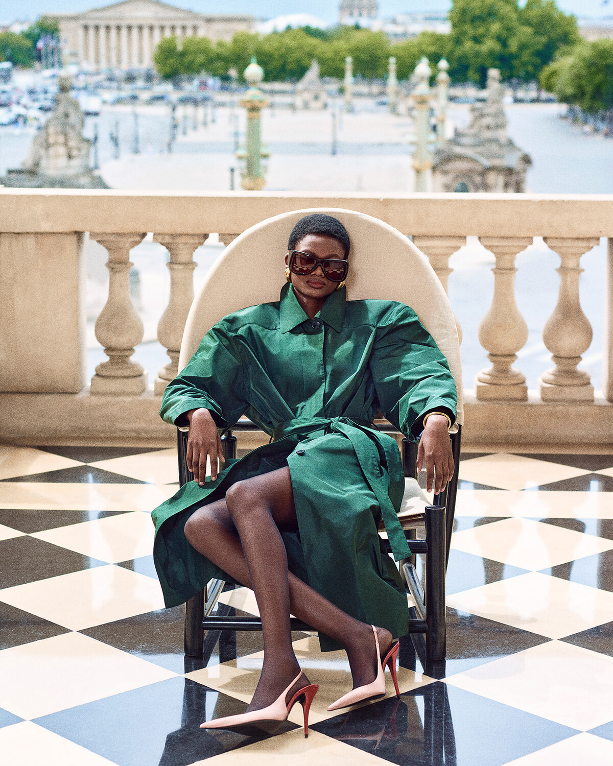 Woman in an emerald trench coat and pink heels sits in a chair on a black-and-white checkered terrace, wearing oversized sunglasses.