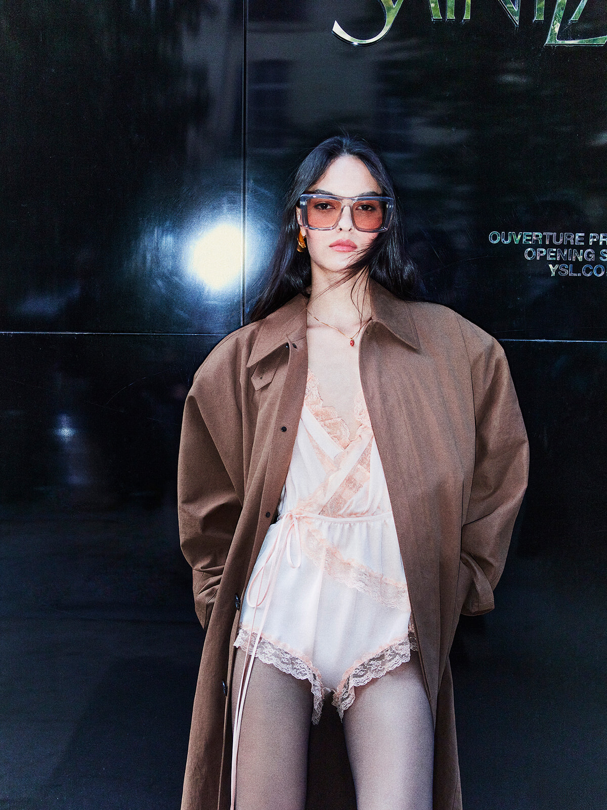 Model posing in an oversized brown coat over pink lace lingerie with pink-tinted sunglasses against a dark glossy wall.