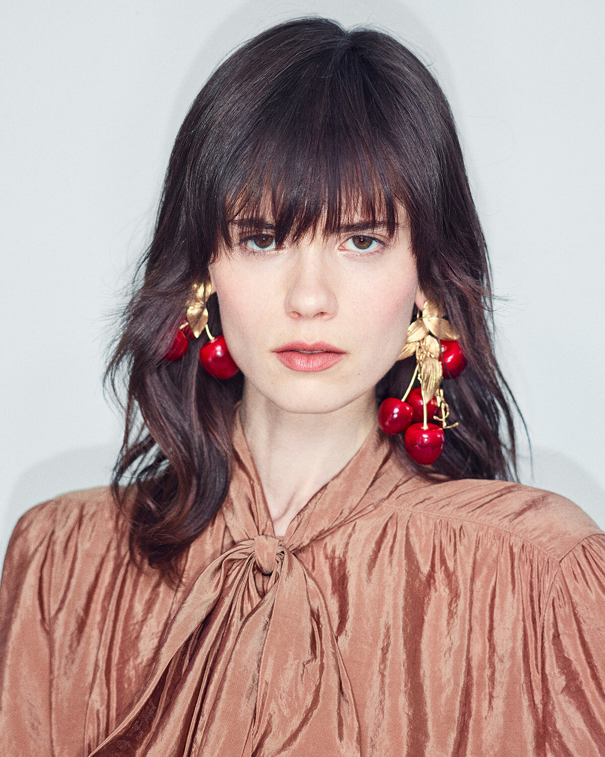 Portrait of a woman with dark hair and cherry earrings, wearing a bronze satin blouse with a tied neck
