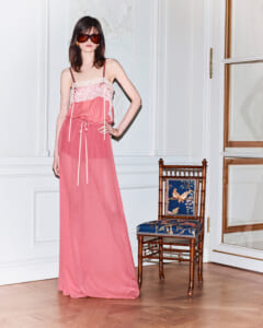 Model in pink lace-trim spaghetti-strap maxi dress with oversized sunglasses posing in a white-paneled room with a wooden chair nearby.